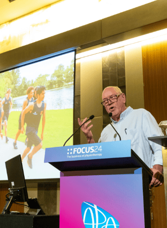 A man is leaning on the podium as he gives a conference presentation. IN the background the screen shows Indigenous Australian boys training for sport.. 